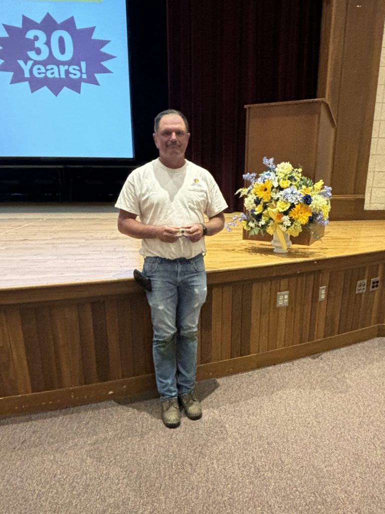 A man poses in front of a stage holding a service pin.