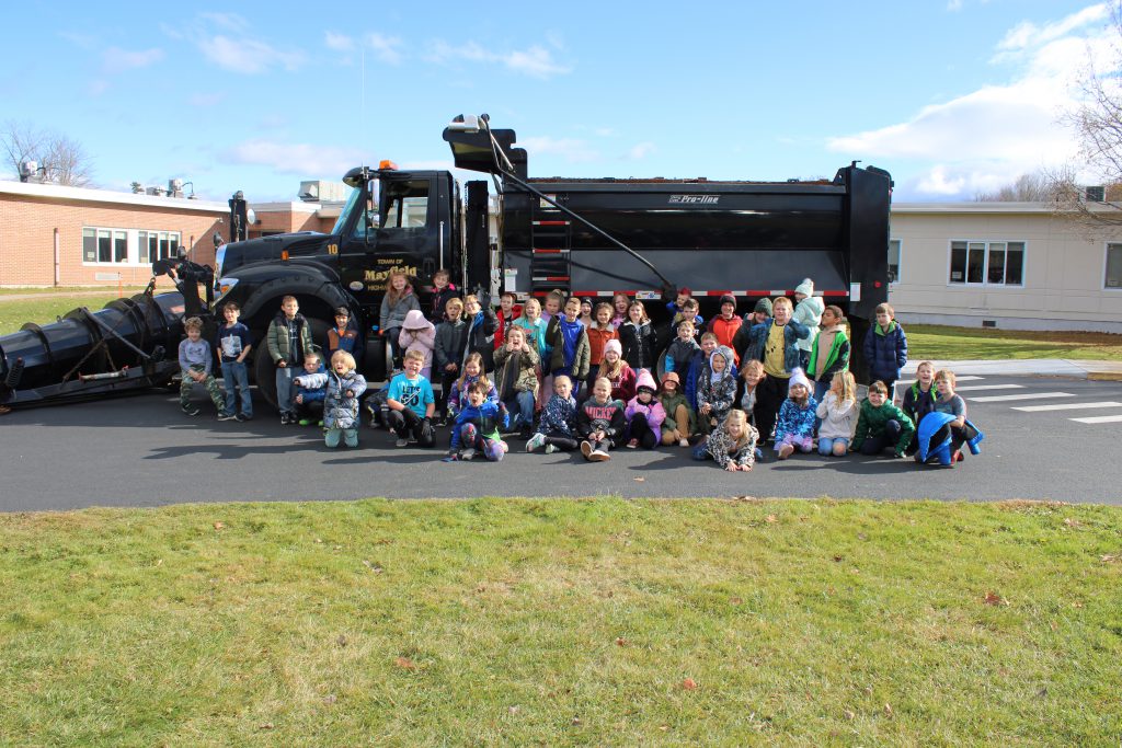A class of young students smiles in front of a black Town of Mayfield dump truck