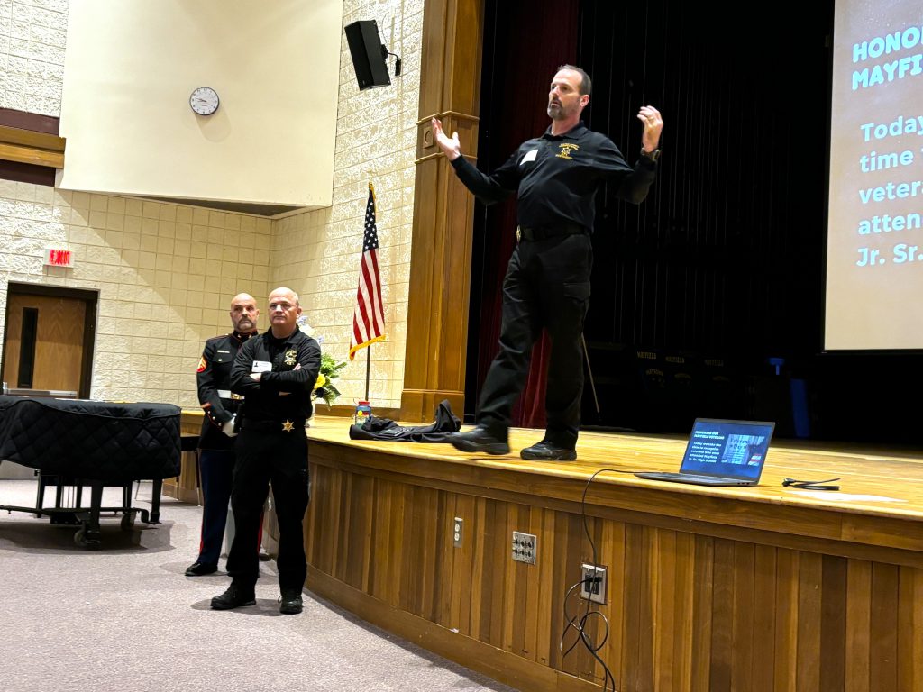 Man stands on stage with his hands up as he talked. Two other men in uniform stand on the floor below.