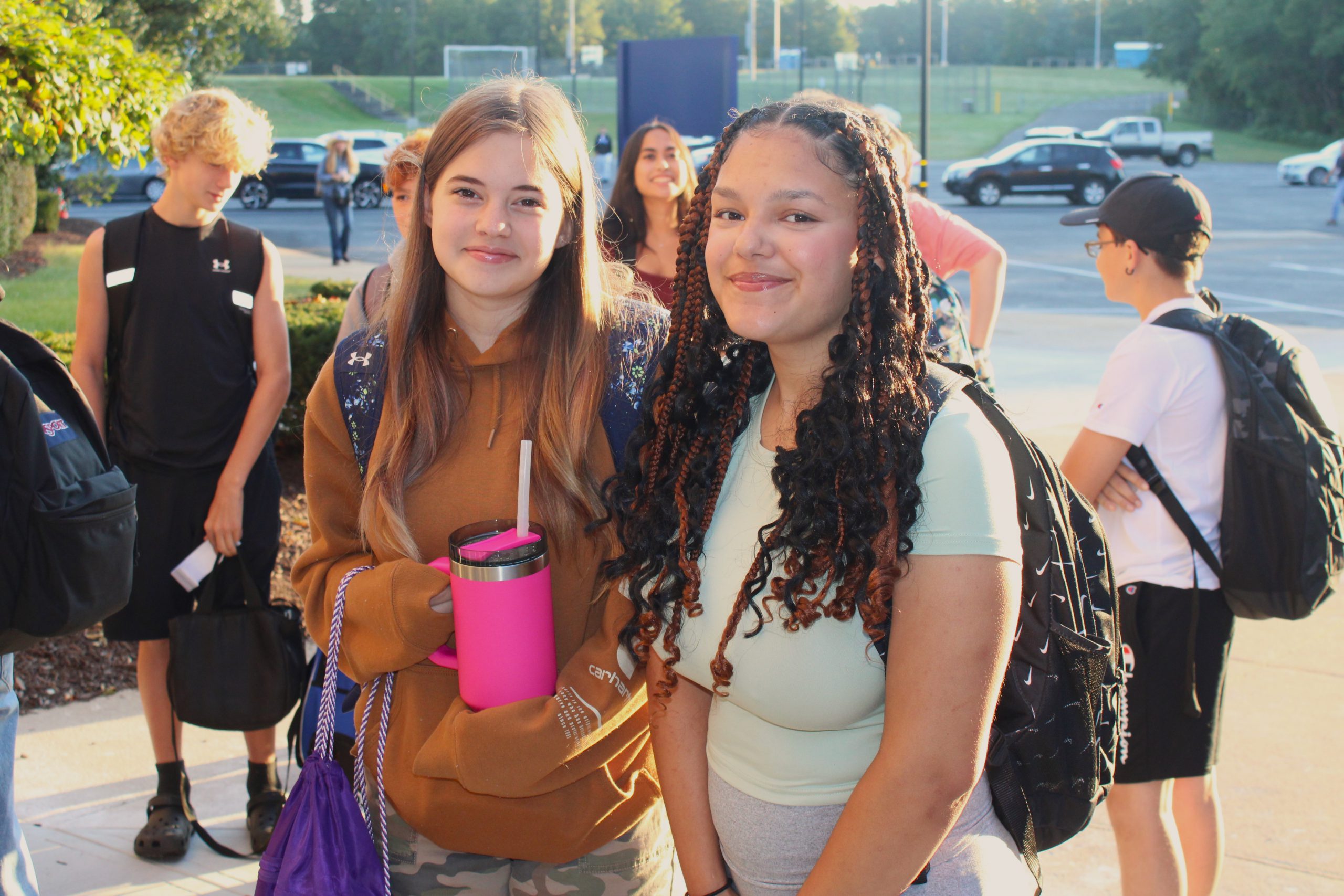 two students pose outside the school wearing bookbags while other students stand behind