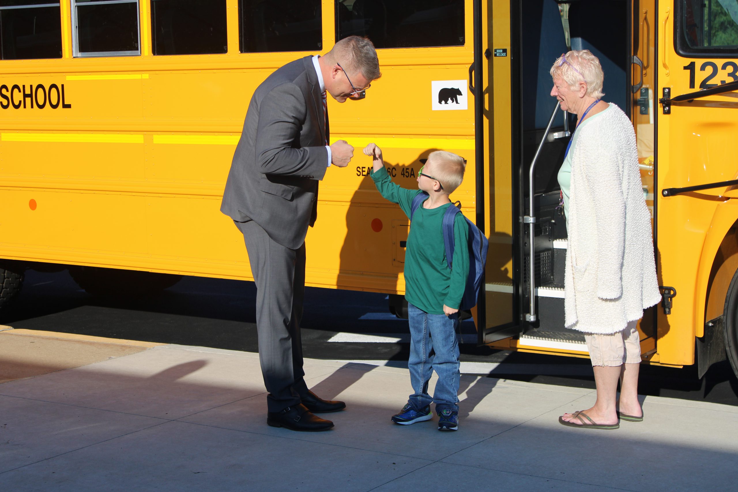 man in suit fist bumps small boy in front of a school bus while a woman looks on and smiles