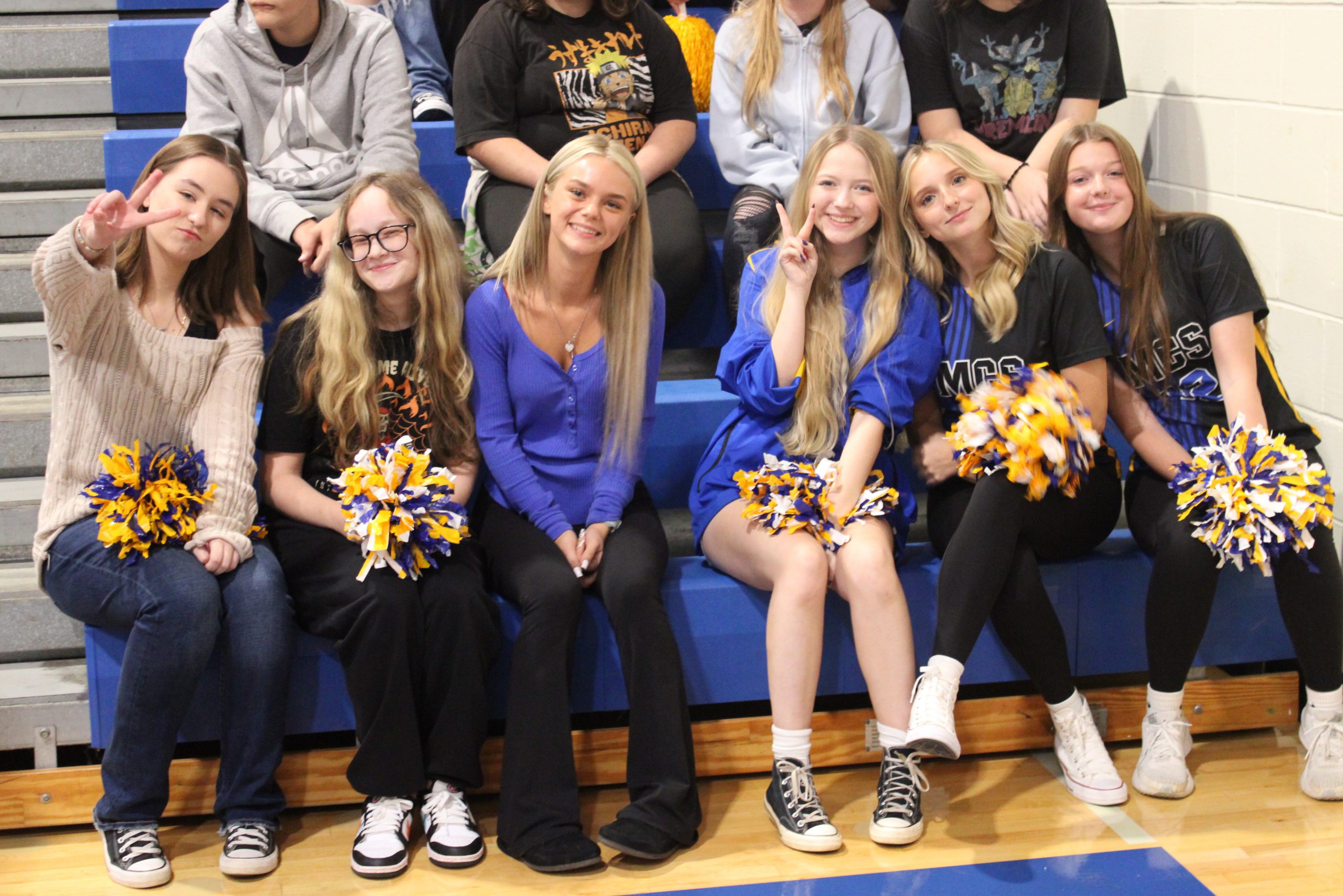 six students hold pompoms and pose on the bleachers