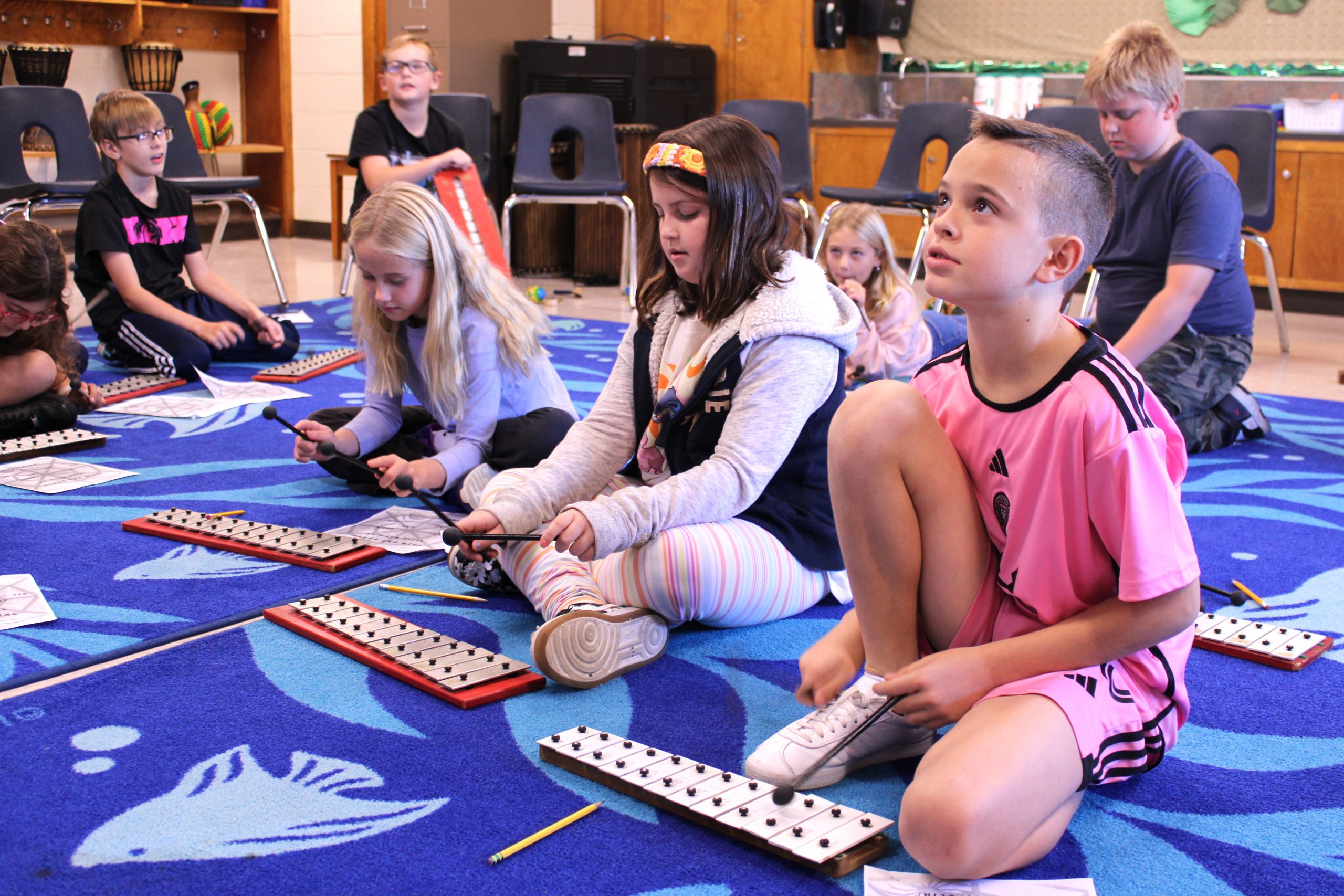 students sit on the carpet in a music room and listen to the teacher while playing instruments